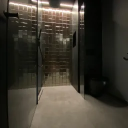 Nowoczesna łazienka w ciemnych kolorach z kabiną prysznicową wyłożoną mozaiką, oświetleniem LED i czarną toaletą.