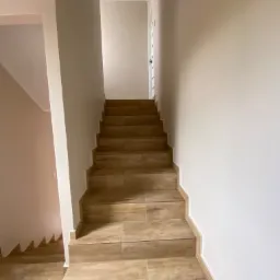 Schody wewnętrzne wyłożone płytkami imitującymi drewno, prowadzące na g&oacute;rę, z widoczną białą ścianą po prawej stronie i fragmentem ściany w odcieniach beżu po lewej stronie.