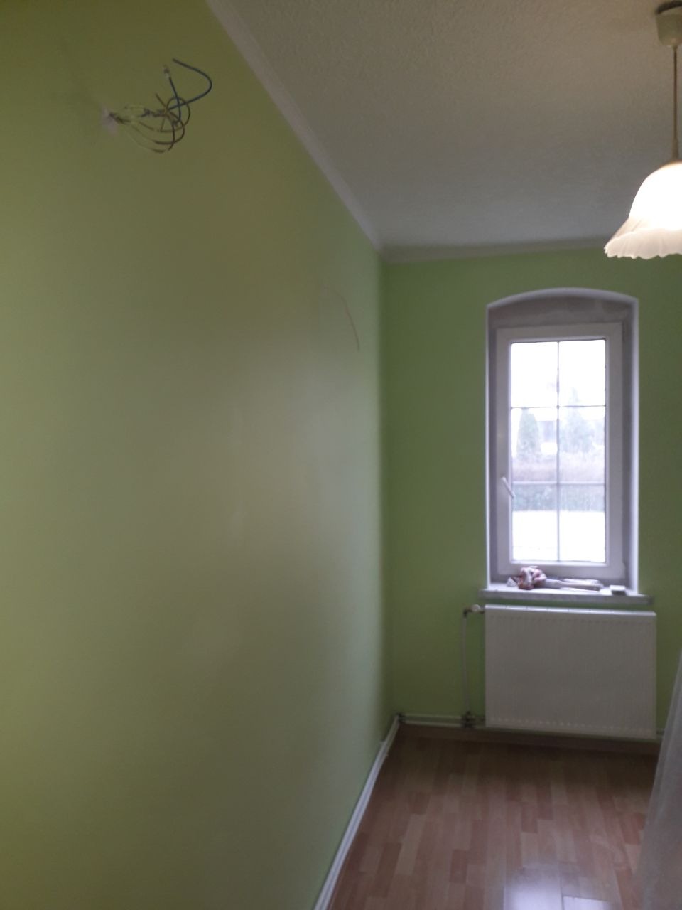 Pokój w odcieniach zieleni z drewnianą podłogą, grzejnikiem pod oknem i wystającymi kablami ze ściany, czekający na wykończenie.