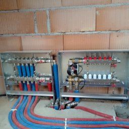 EUkalipt - Rozdzielacze ogrzewania podłogowego w metalowych obudowach, z przewodami czerwono-niebieskimi, zamontowane na tle ściany z cegły ceramicznej.