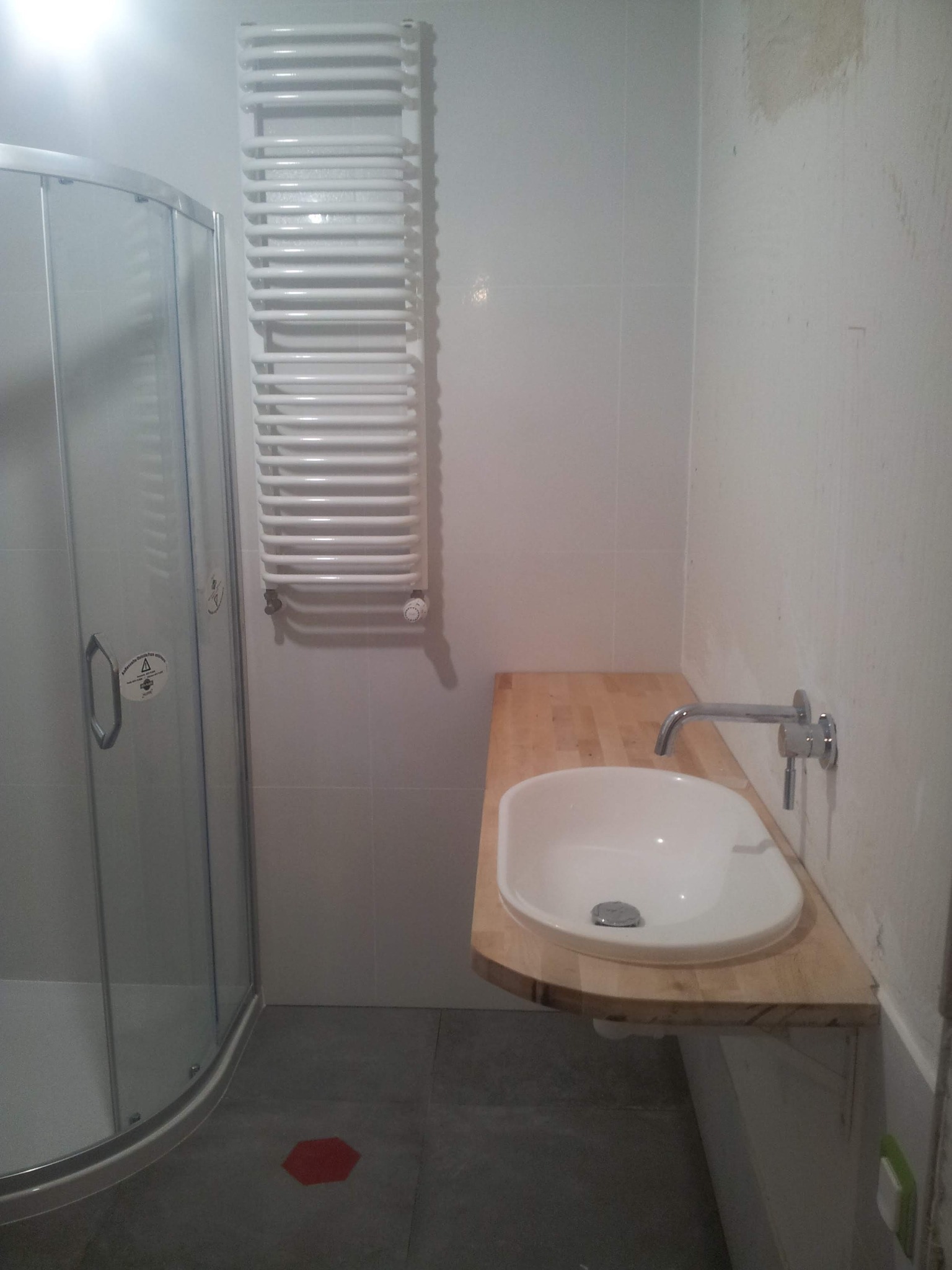 Nowoczesna łazienka z kabiną prysznicową, grzejnikiem drabinkowym i umywalką nablatową na drewnianym blacie, widoczne ślady prac wykończeniowych na ścianie.