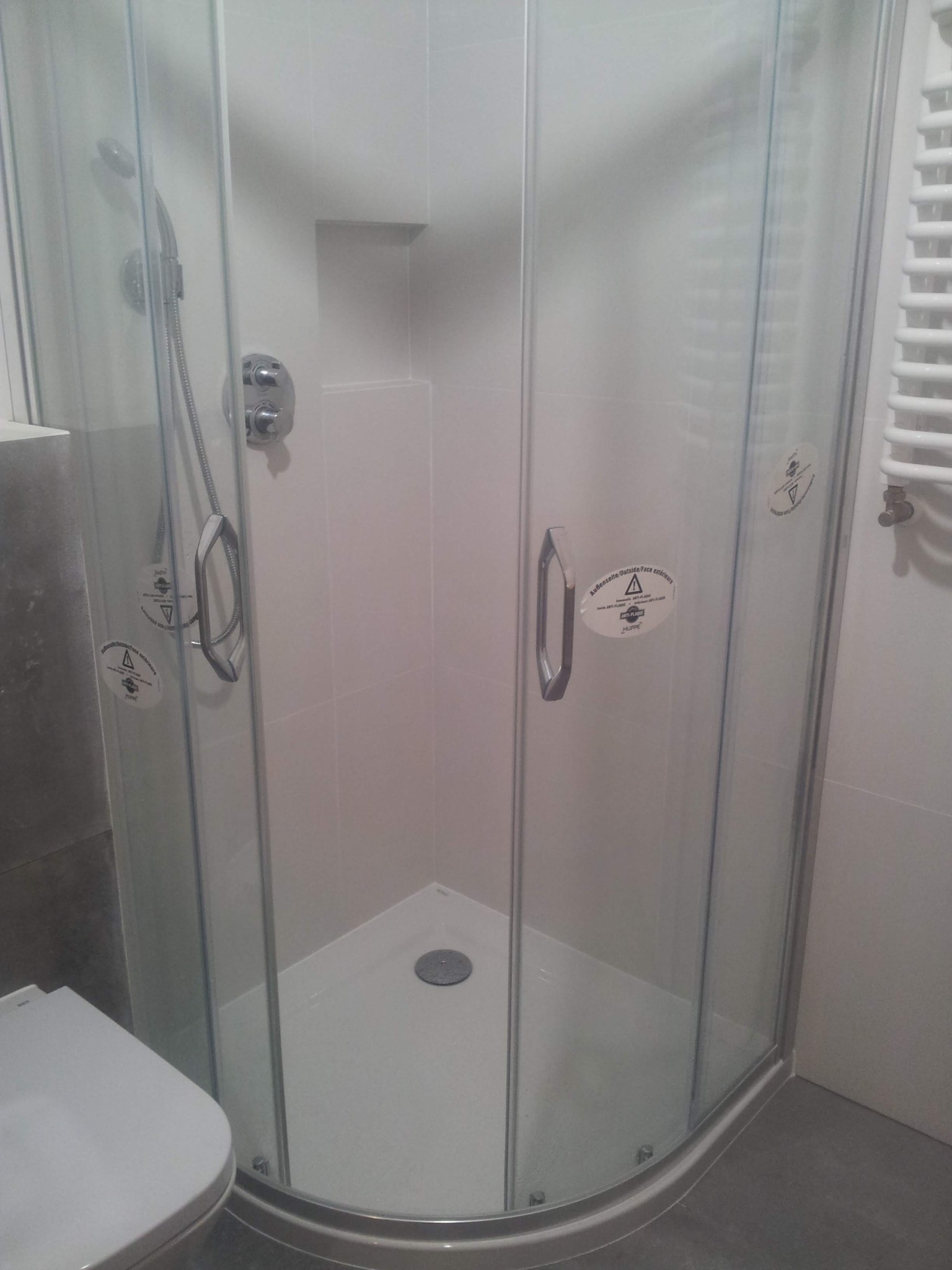 Narożna kabina prysznicowa z przezroczystego szkła z chromowaną armaturą i naklejkami ostrzegawczymi na szklanych drzwiach, widoczny fragment toalety w tle.