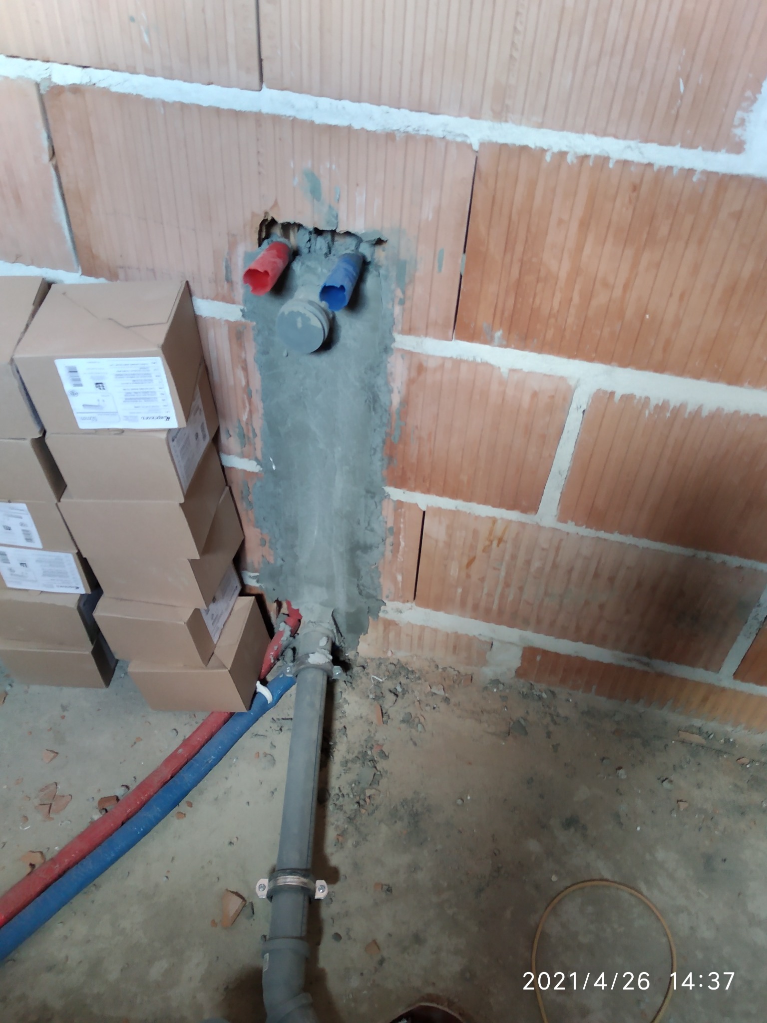 Wykute w ceglanej ścianie otwory na instalację wodną z wystającymi rurami w kolorze niebieskim i czerwonym, zalane szarą zaprawą, poniżej pionowa rura kanalizacyjna z obejmami, na podłodze gruz...