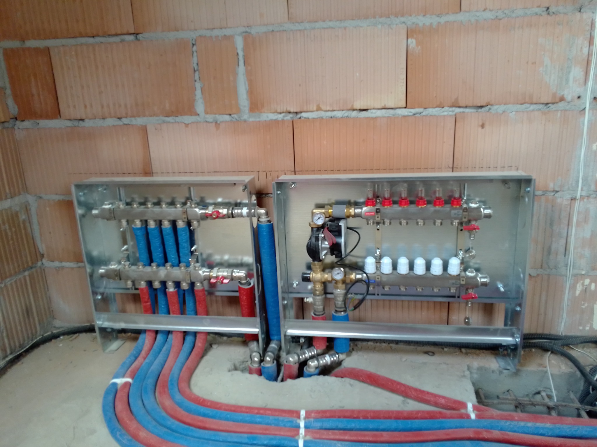 Rozdzielacze ogrzewania podłogowego w metalowych obudowach, z przewodami czerwono-niebieskimi, zamontowane na tle ściany z cegły ceramicznej.