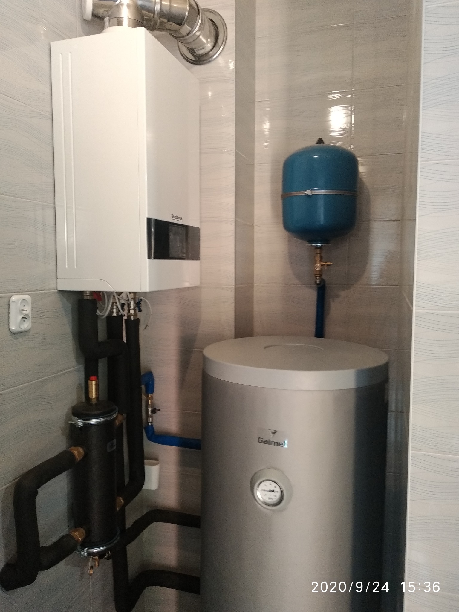 Biały piec gazowy Buderus z podłączeniami, pionowy srebrny zasobnik Galmet z termometrem i niebieskie naczynie przeponowe w kotłowni z szarymi płytkami.