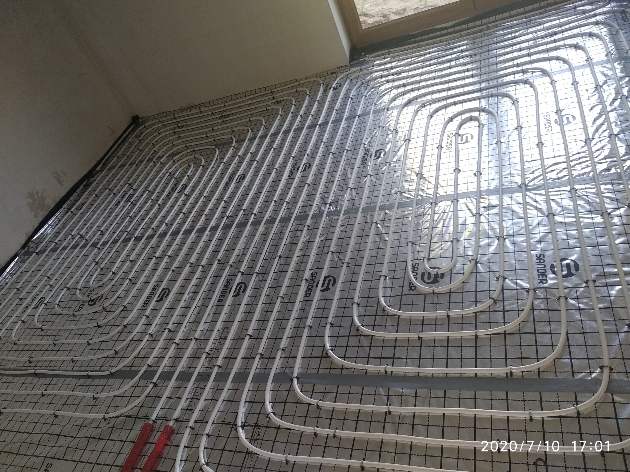 Białe rury grzewcze ułożone w pętle na podłodze, zamocowane do czarnej siatki na srebrnej folii izolacyjnej, widoczne czerwone elementy mocujące.