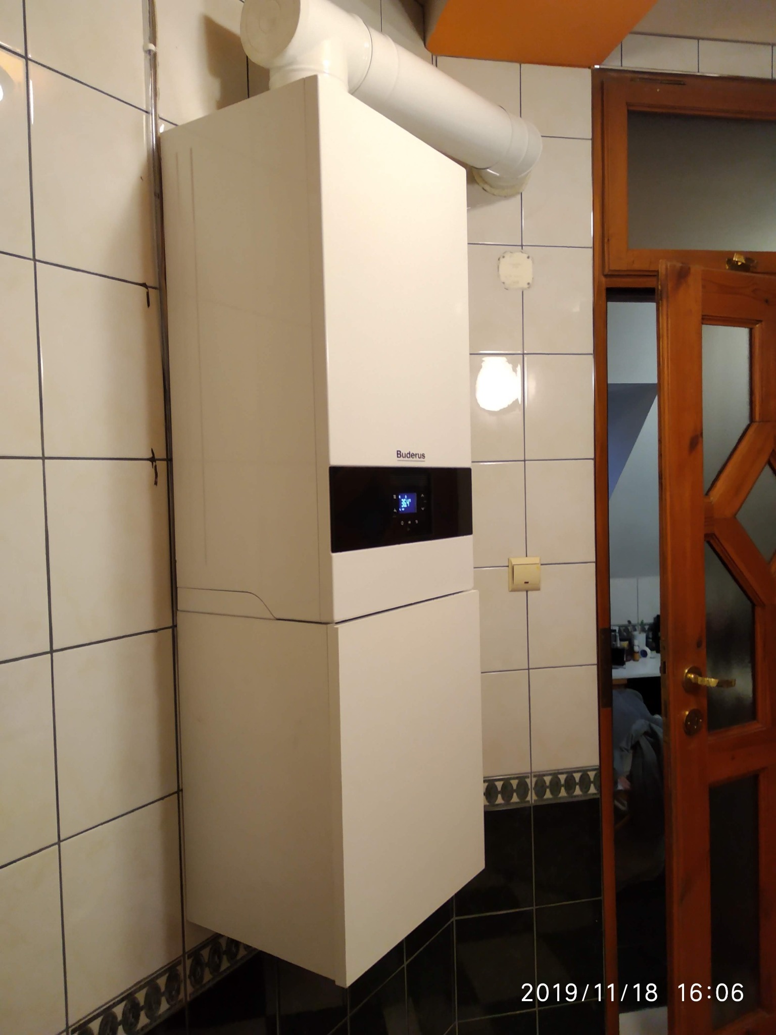 Biały, wiszący piec gazowy marki Buderus z czarnym panelem sterowania, zamontowany na kafelkowanej ścianie w łazience, widoczny fragment drewnianych drzwi.
