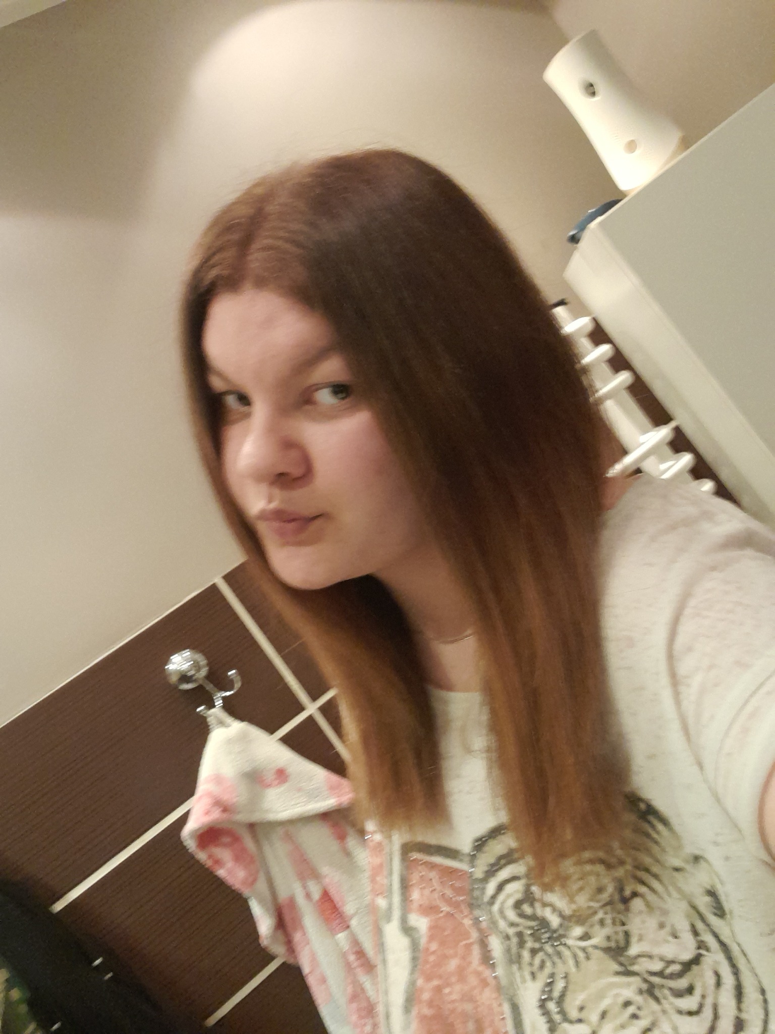 Portret kobiety z prostymi, brązowymi włosami, wykonany w łazience z widocznym ręcznikiem na wieszaku i dozownikiem odświeżacza powietrza w tle.