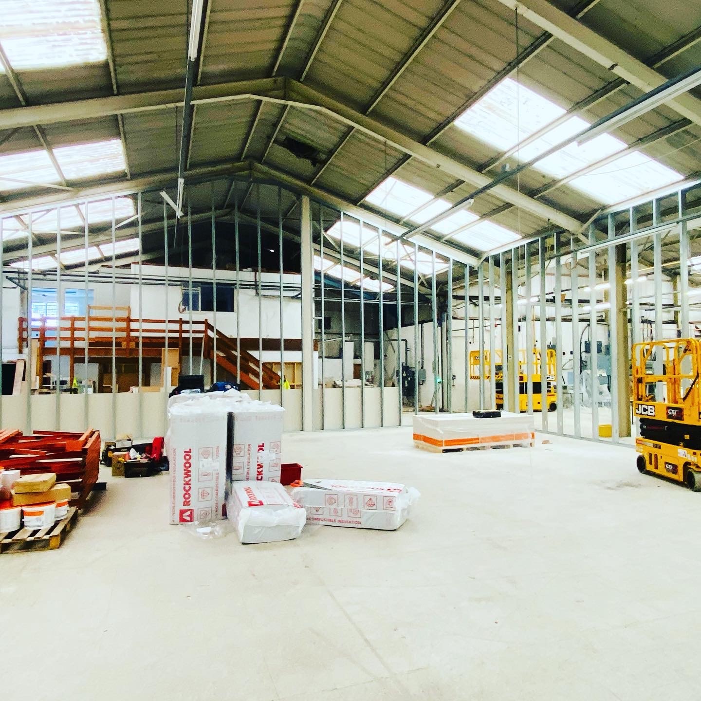 Wnętrze hali w trakcie budowy ścian działowych z metalowych profili, widoczne pakiety wełny mineralnej Rockwool, podnośnik nożycowy JCB oraz elementy konstrukcji drewnianej w tle.