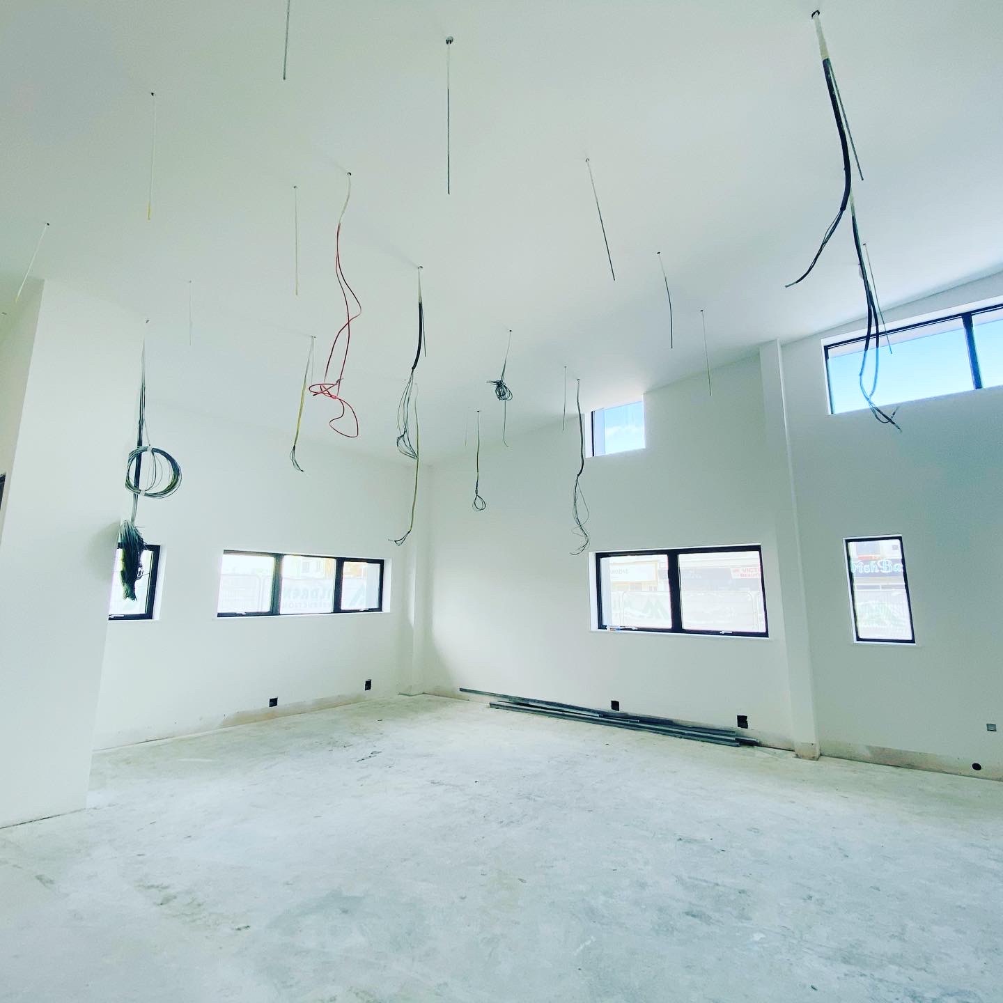 Wnętrze w trakcie prac wykończeniowych, białe ściany, surowa podłoga, wiszące przewody elektryczne i profile pod sufitem, okna w czarnych ramach.