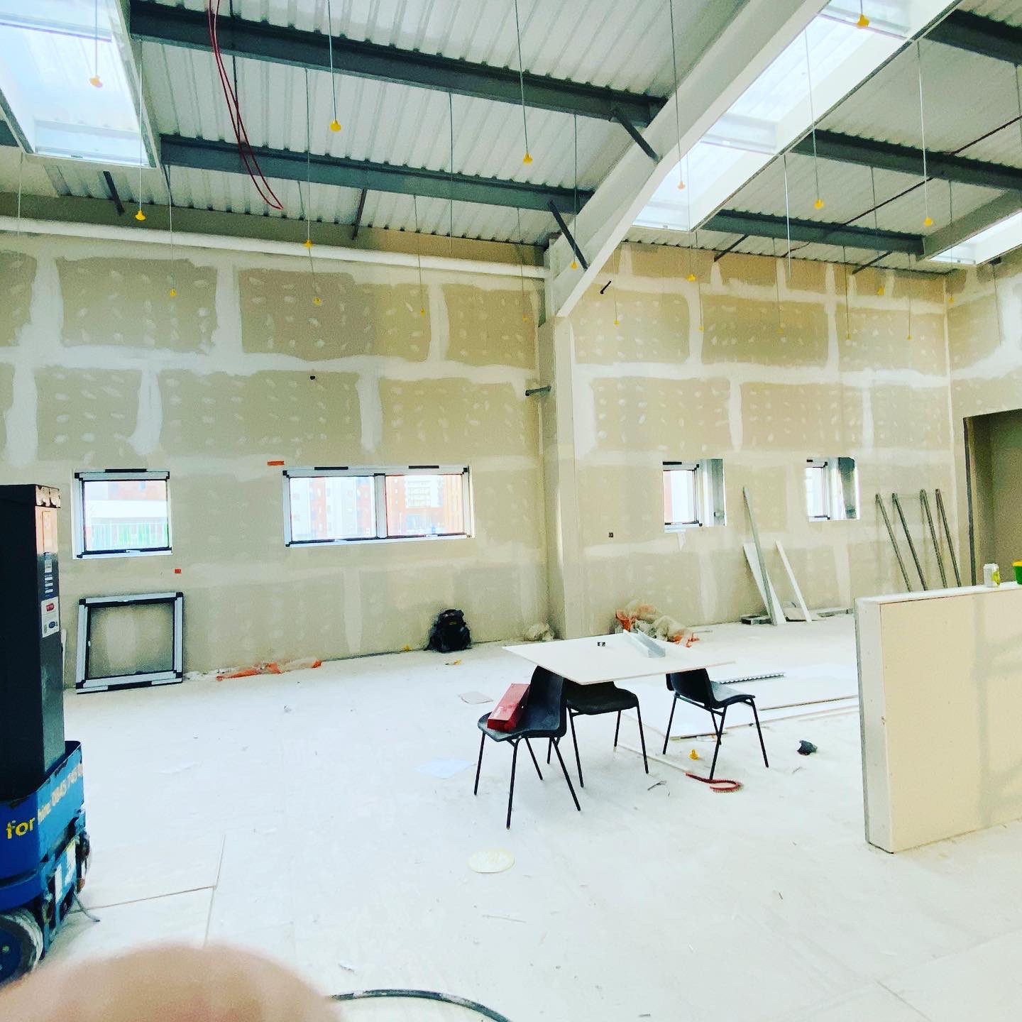 Wnętrze biurowe w trakcie prac wykończeniowych: ściany z płyt kartonowo-gipsowych po szpachlowaniu, widoczne okna, metalowa konstrukcja sufitu z oświetleniem, tymczasowe meble i narzędzia.