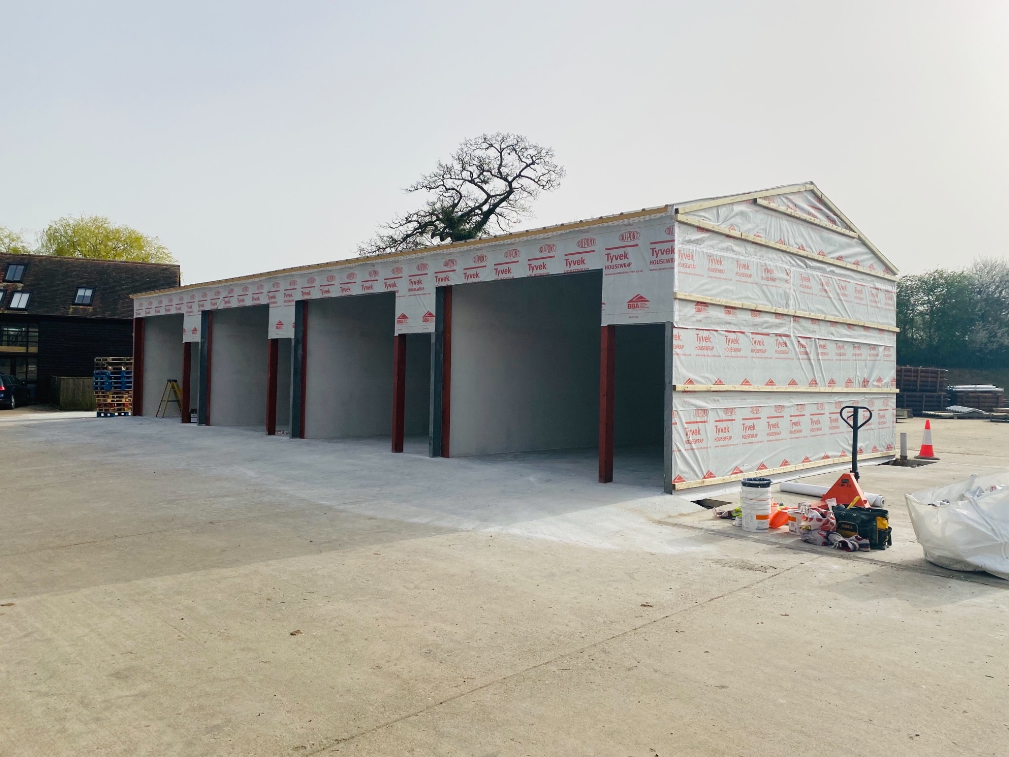 Budynek w trakcie budowy z widoczną izolacją Tyvek, surowym betonem i elementami konstrukcyjnymi, paleta z materiałami budowlanymi w tle.