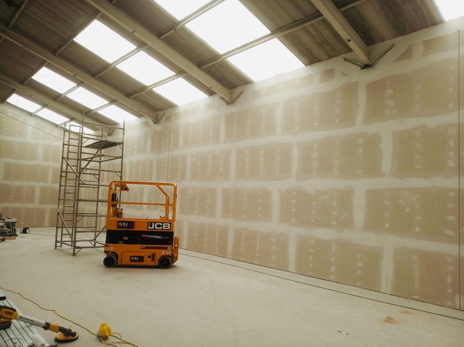 Duża hala z ścianą pokrytą płytami kartonowo-gipsowymi przed szpachlowaniem, widoczny podnośnik nożycowy JCB i rusztowanie.