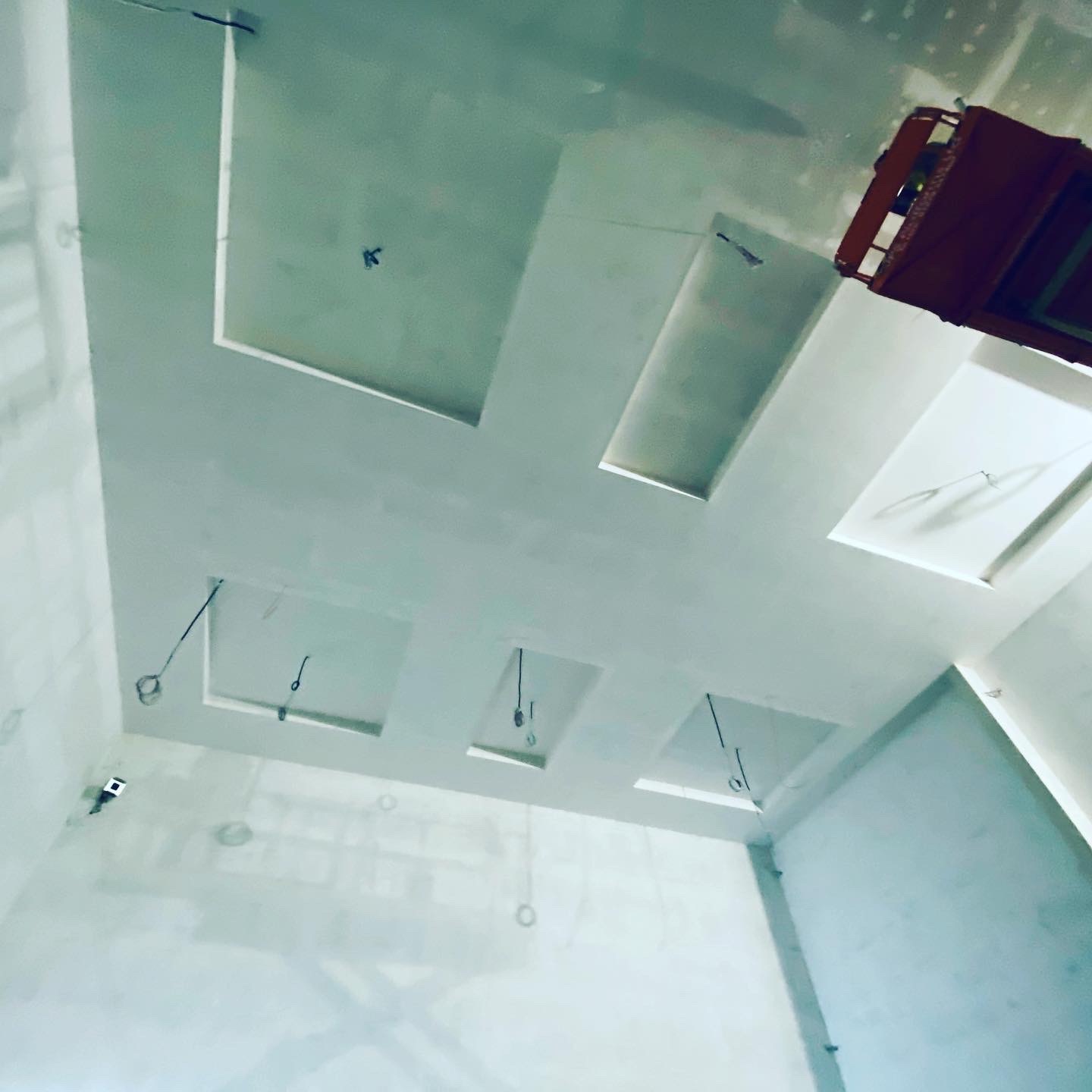 Wnętrze w trakcie prac wykończeniowych, sufit z wnękami oświetleniowymi w kształcie kwadratów i prostokątów, widoczne przewody elektryczne zwisające z sufitu, ściany po szpachlowaniu, w rogu...