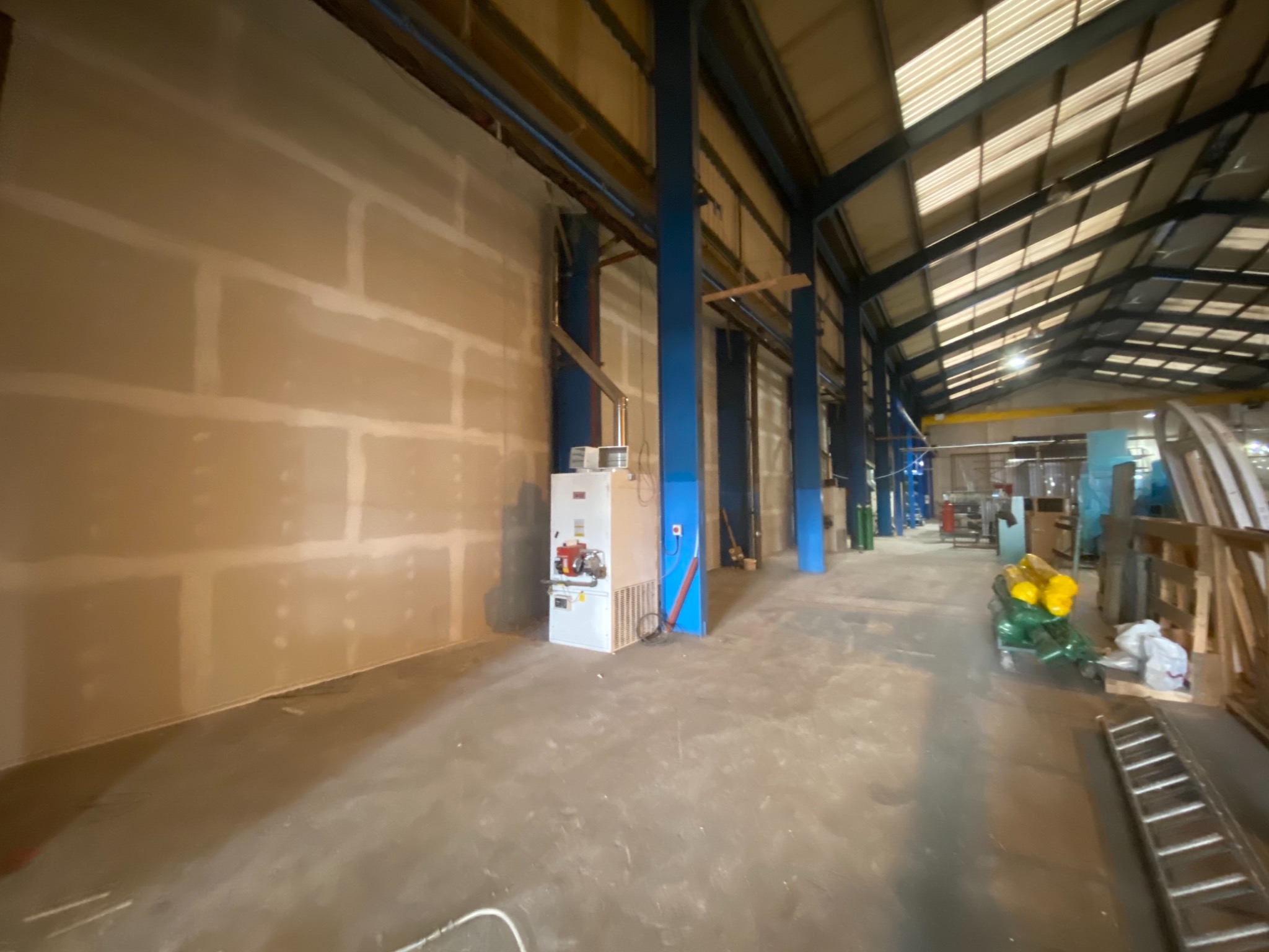 Wnętrze hali w trakcie prac wykończeniowych; ściana z płyt gipsowo-kartonowych przed szpachlowaniem, widoczne łączenia płyt; w tle elementy konstrukcyjne hali, drabina i materiały budowlane.