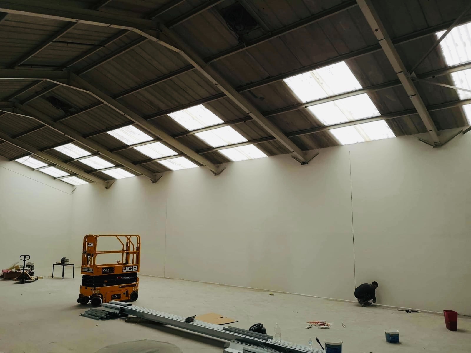 Wnętrze hali przemysłowej w trakcie prac wykończeniowych; widoczna podnośnik koszowy JCB, pracownik przy listwie przypodłogowej oraz sufit z oknami dachowymi.