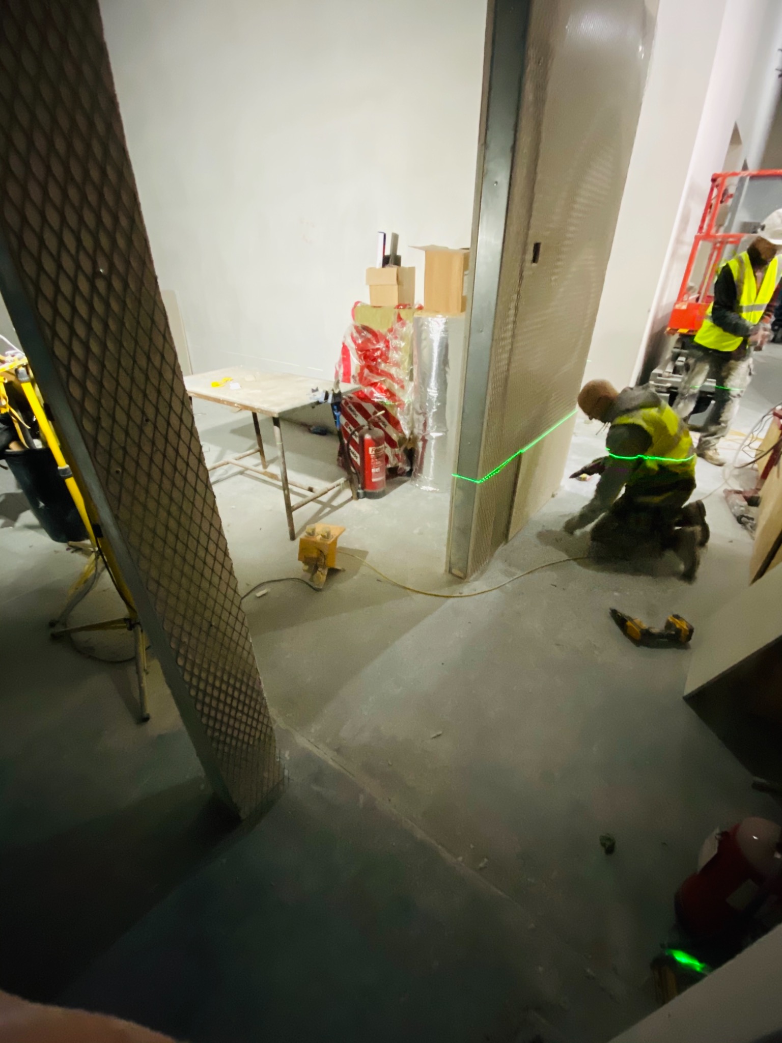 Montaż paneli ściennych z użyciem lasera krzyżowego, widoczny pracownik w kamizelce odblaskowej na kolanach, drugi pracownik na podnośniku koszowym w tle, narzędzia i materiały budowlane...