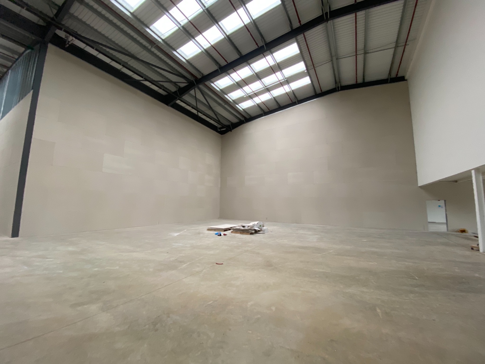 Wnętrze hali przemysłowej w trakcie prac wykończeniowych, ściany pokryte płytami kartonowo-gipsowymi, widoczne materiały budowlane na podłodze, sufit z licznymi świetlikami.