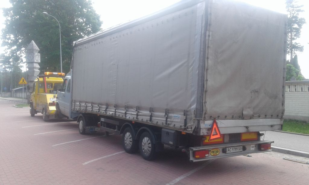 Żółta laweta holuje szarą ciężarówkę z plandeką na parkingu wyłożonym czerwoną kostką brukową. W tle drzewa i pomnik.