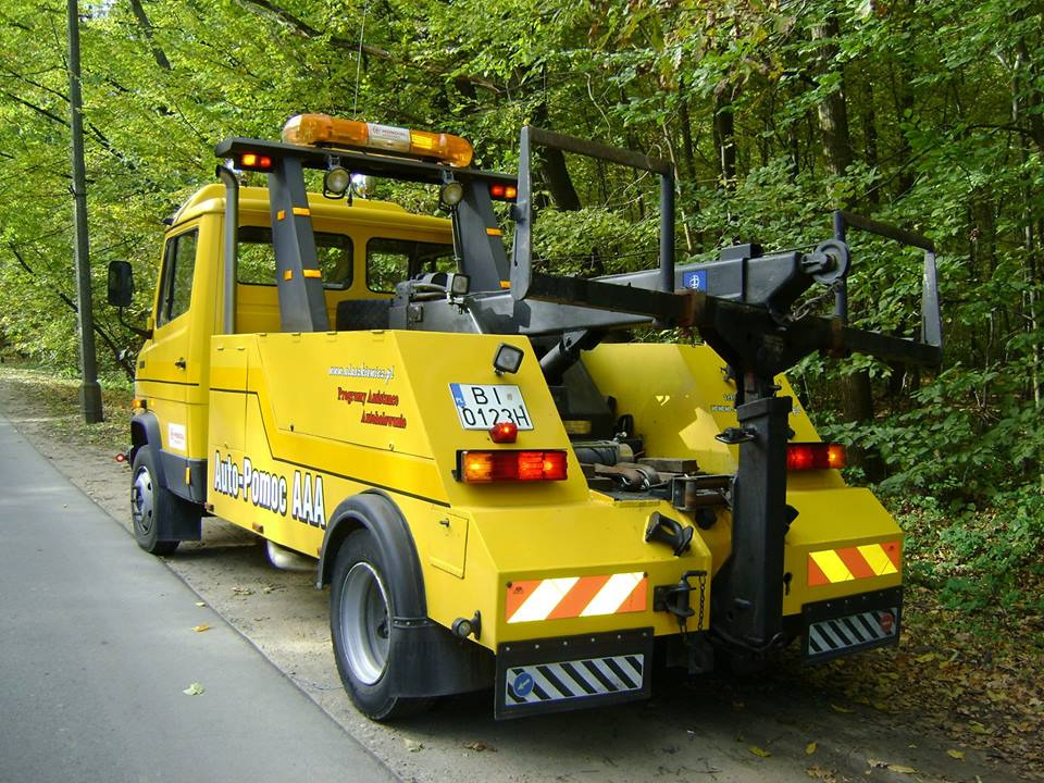 Żółta laweta Auto-Pomoc AAA zaparkowana na poboczu drogi w lesie, widok z tyłu, włączone światła ostrzegawcze.