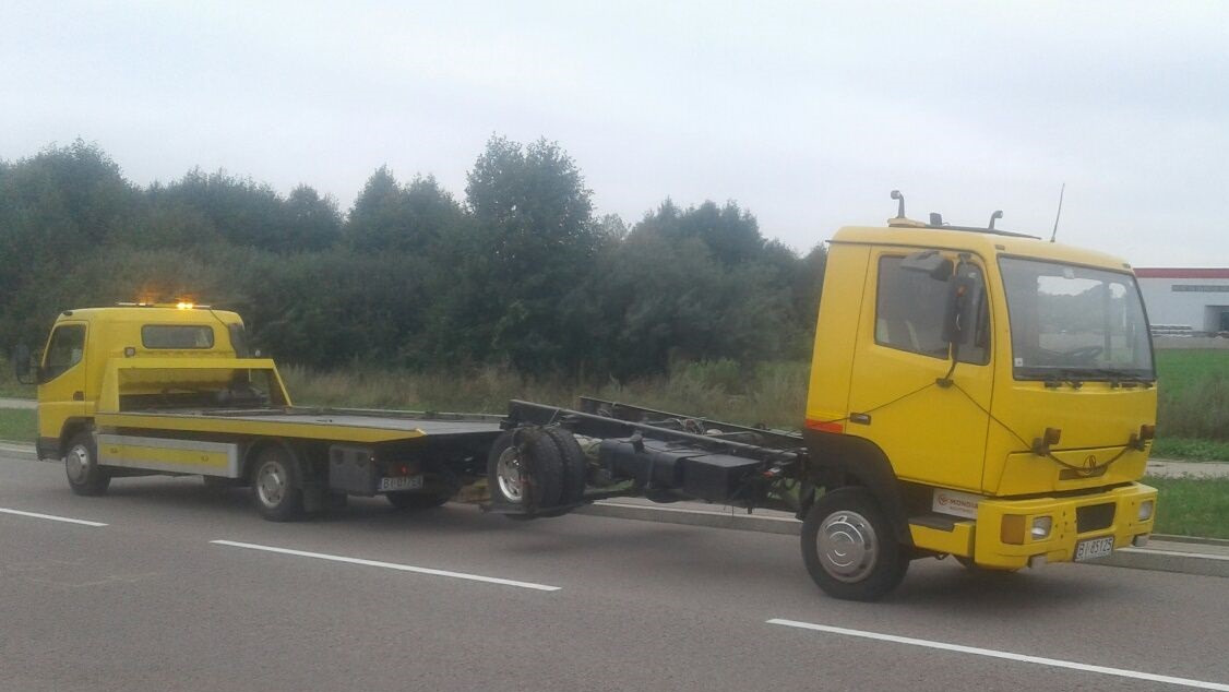 Żółta laweta z włączonymi światłami ostrzegawczymi stoi na poboczu drogi, widoczna tablica rejestracyjna, z tyłu las i budynek.