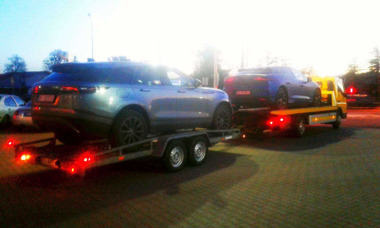 Dwa samochody, srebrny SUV i niebieskie sportowe coupe, transportowane na żółtej lawecie z włączonymi światłami ostrzegawczymi, na tle miejskiego otoczenia o zmierzchu.