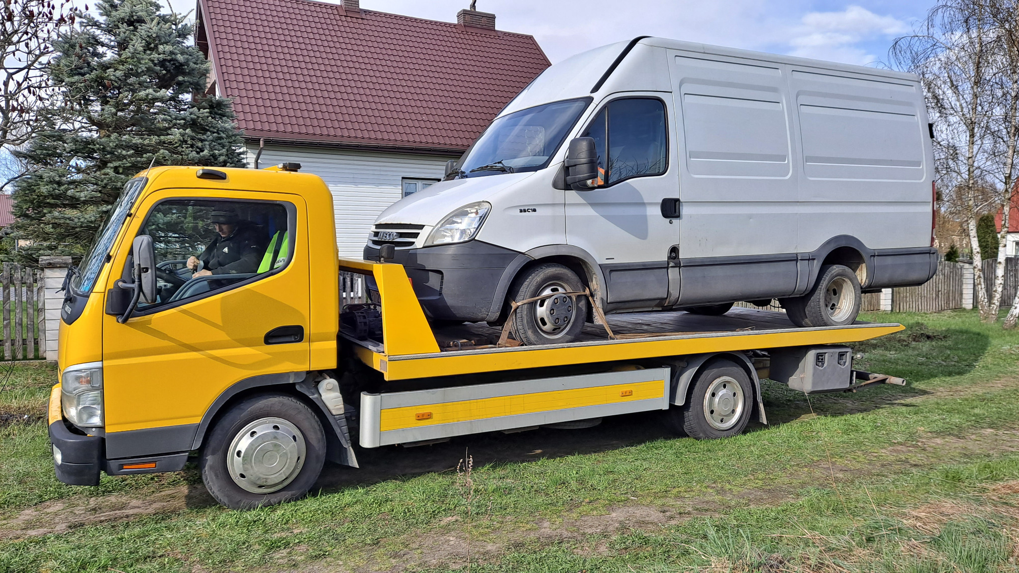 Żółta laweta z białym busem Iveco Daily zabezpieczonym pasami, na tle domu z czerwoną dachówką i zielonego trawnika.