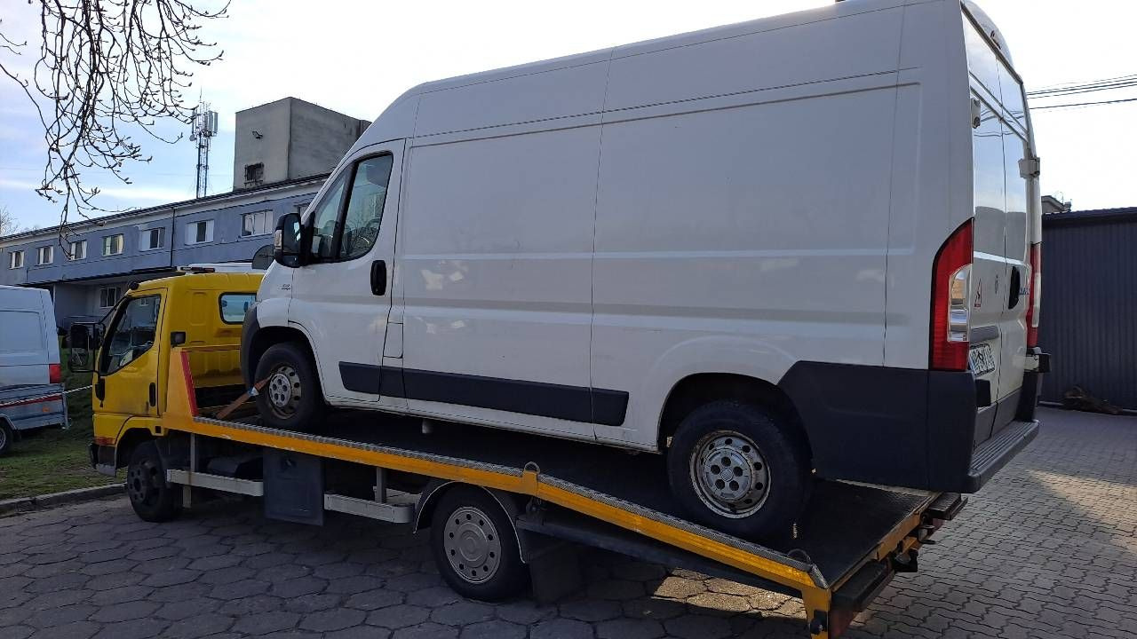 Żółta laweta przewozi białego busa na tle szarego budynku z anteną telefonii komórkowej.