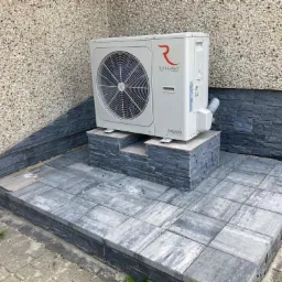 Biała jednostka zewnętrzna z wentylatorem marki Rotenso Aquami Series, umieszczona na podwyższeniu z szarych płyt chodnikowych i kamienia łupanego, na tle ściany budynku z elewacją z kamyczków.