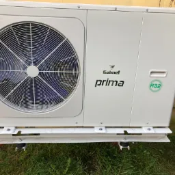 Zewnętrzna jednostka urządzenia grzewczego Galmet Prima R32 zamontowana na metalowej konstrukcji przy żółtej ścianie budynku. Widoczny wentylator i logo producenta.