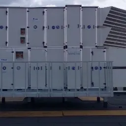 Rozbudowana instalacja rekuperacyjna na dachu budynku, widoczne segmenty z logo producenta i kratki wentylacyjne, tło stanowi zachmurzone niebo i drzewa.