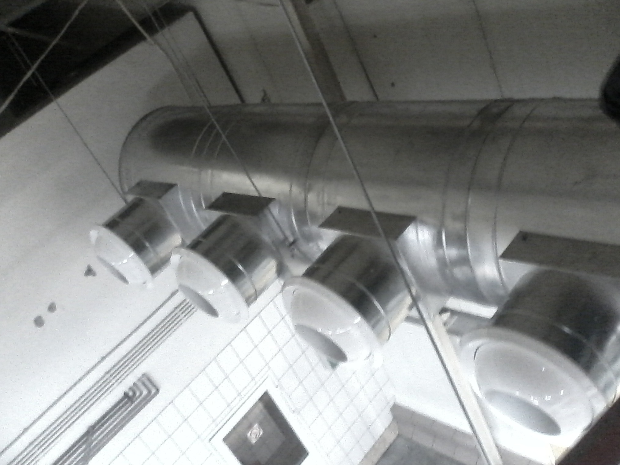 Metalowy system wentylacyjny z okrągłymi wylotami zamontowany pod sufitem z białych płytek, widoczne przewody i elementy konstrukcyjne.