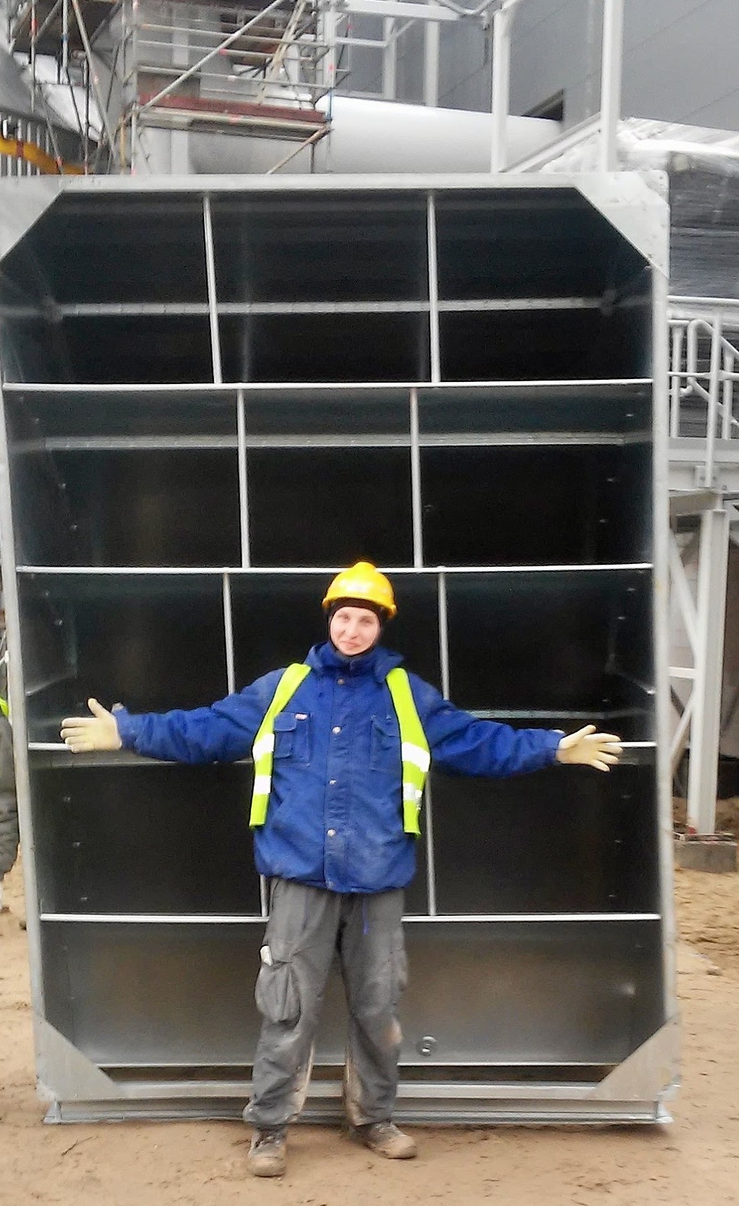 Pracownik w kasku i kamizelce odblaskowej stoi przed dużym, metalowym modułem rekuperacyjnym z wewnętrznymi przegrodami, na placu budowy.