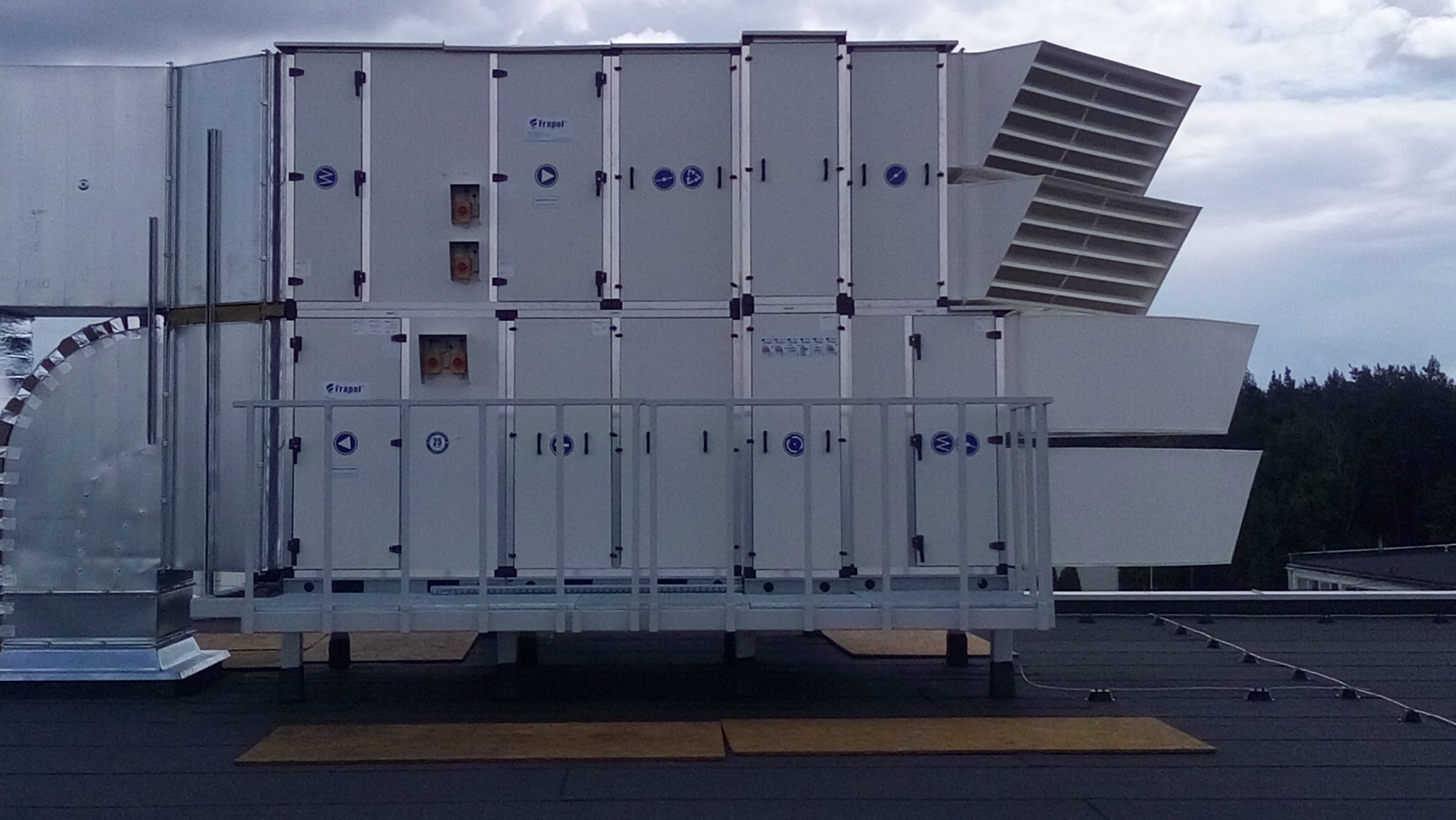 Rozbudowana instalacja rekuperacyjna na dachu budynku, widoczne segmenty z logo producenta i kratki wentylacyjne, tło stanowi zachmurzone niebo i drzewa.