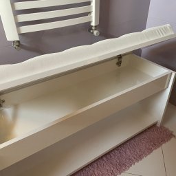 O L B - Biała szafka na buty z tapicerowanym siedziskiem, otwierana klapa, widoczny mechanizm podtrzymujący, na tle fioletowych płytek i grzejnika drabinkowego.