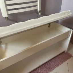 Biała szafka na buty z tapicerowanym siedziskiem, otwierana klapa, widoczny mechanizm podtrzymujący, na tle fioletowych płytek i grzejnika drabinkowego.