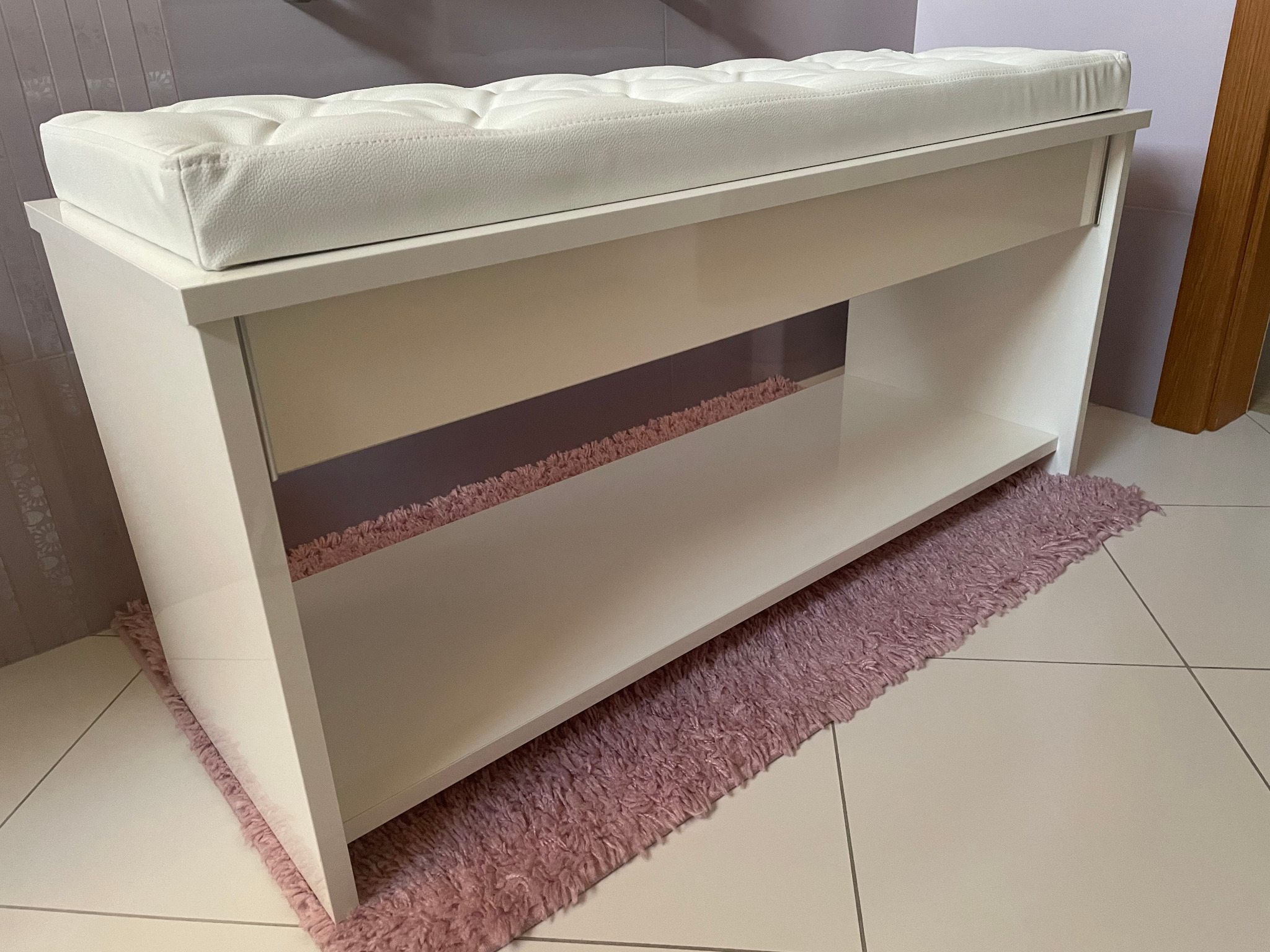 Biała, lakierowana ławeczka z pikowanym siedziskiem z ekoskóry, na tle jasnych płytek podłogowych i puszystego, różowego dywanika.