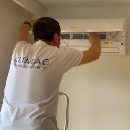 Mężczyzna na drabinie, w koszulce firmowej KLIMAG, czyści filtry wewnątrz otwartej, białej jednostki klimatyzacyjnej zamontowanej na ścianie.