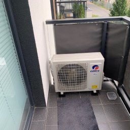 Klimag - Jednostka zewnętrzna klimatyzatora Gree zamontowana na balkonie z szarymi płytkami, widoczna osłona balkonowa i okno z żaluzjami.