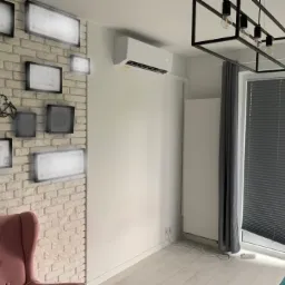 Nowoczesny salon z zamontowaną białą jednostką klimatyzacyjną na ścianie, widoczna ceglana ściana z ramkami, designerska lampa sufitowa i szare rolety okienne.