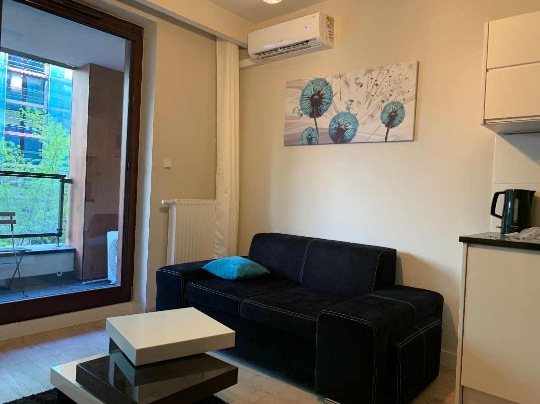 Nowoczesny klimatyzator ścienny zamontowany w jasnym salonie z balkonem i czarną sofą, widoczny na tle obrazu z dmuchawcami.