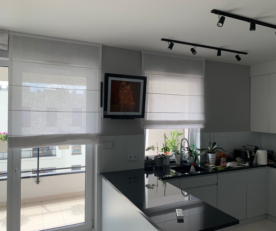 Nowoczesna kuchnia z białymi roletami rzymskimi, czarnym blatem, oświetleniem punktowym na szynach i telewizorem z obrazem Gustava Klimta na ścianie.