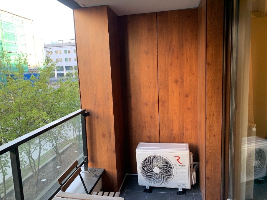 Zewnętrzna jednostka klimatyzatora Rotenso zamontowana na balkonie z drewnianą zabudową, widok na zieleń miejską i budynki w tle.