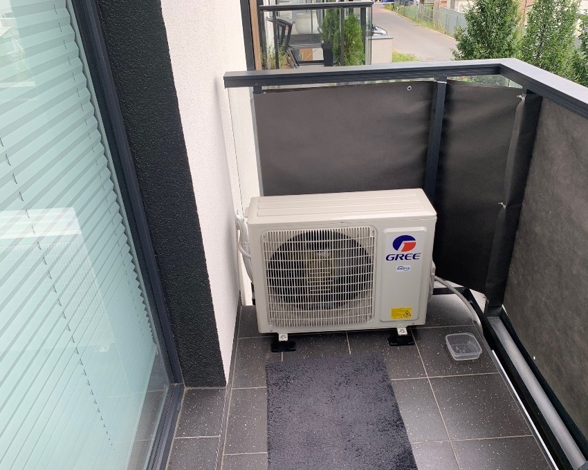 Jednostka zewnętrzna klimatyzatora Gree zamontowana na balkonie z szarymi płytkami, widoczna osłona balkonowa i okno z żaluzjami.