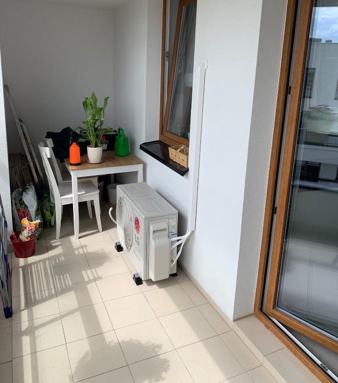 Biała jednostka zewnętrzna klimatyzatora LG umieszczona na balkonie z jasnymi płytkami podłogowymi, widoczne okno i stół z roślinami w tle.