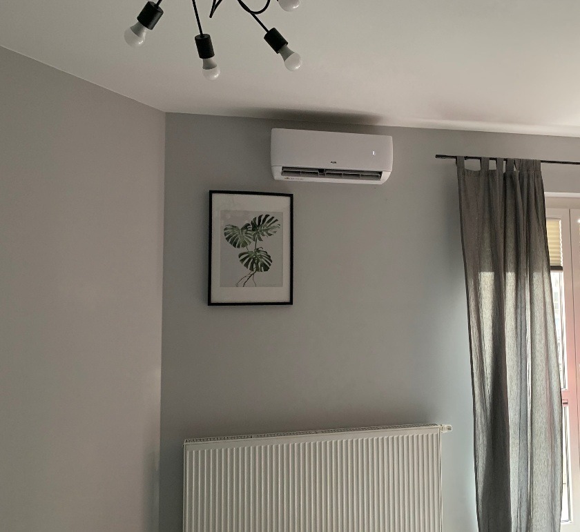Biały klimatyzator ścienny zamontowany nad obrazem z motywem roślinnym w szarej sypialni z grzejnikiem i zasłonami.