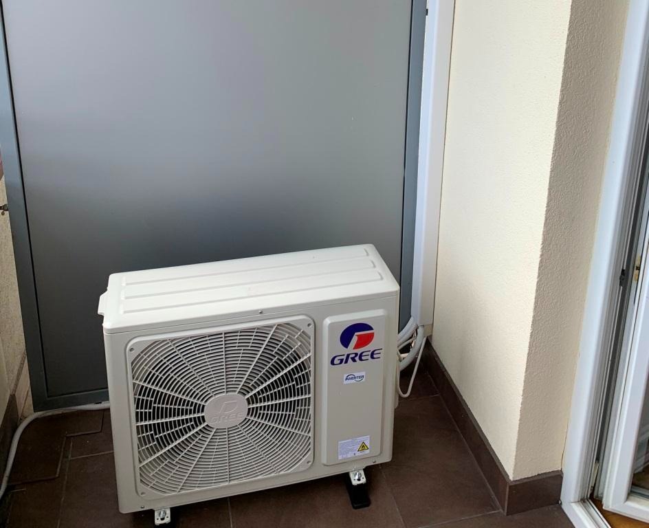 Zewnętrzna jednostka klimatyzacji GREE zamontowana na brązowej podłodze balkonu, widoczne przewody i fragment ściany budynku.