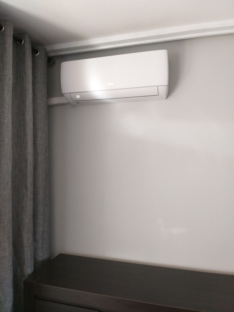 Biały klimatyzator ścienny zamontowany nad ciemnobrązową komodą, obok szarych zasłon w pokoju o jasnych ścianach.