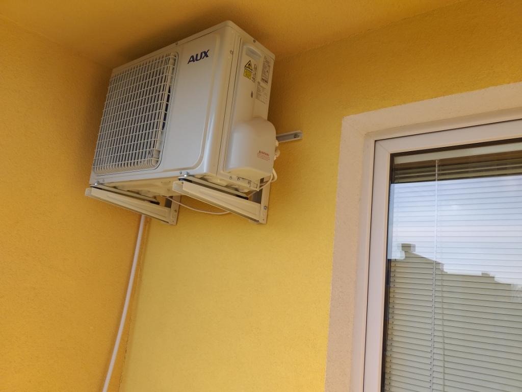 Biała jednostka zewnętrzna klimatyzatora marki AUX zamontowana na żółtej ścianie budynku obok okna z żaluzjami.