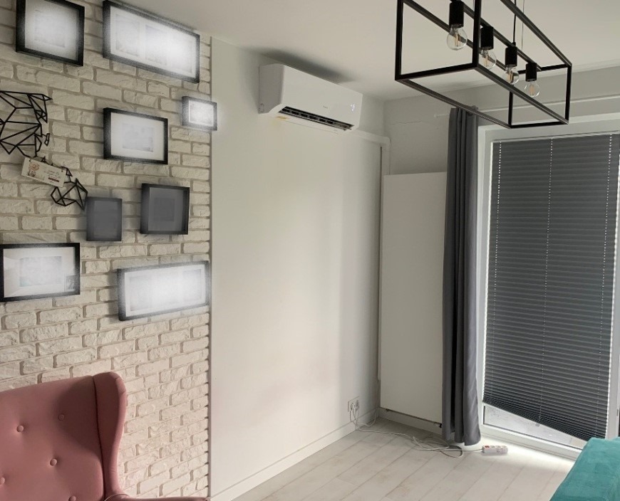 Nowoczesny salon z zamontowaną białą jednostką klimatyzacyjną na ścianie, widoczna ceglana ściana z ramkami, designerska lampa sufitowa i szare rolety okienne.